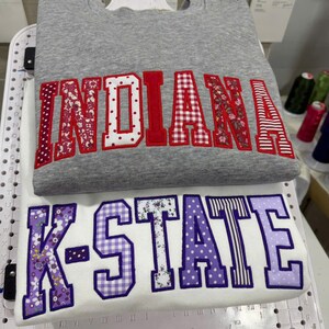 May include: Two sweatshirts, one gray and one white, are stacked. The gray sweatshirt has the word "INDIANA" in red and patterned fabric letters. The white sweatshirt displays "K-STATE" in purple and patterned fabric letters. Both sweatshirts are embroidered.