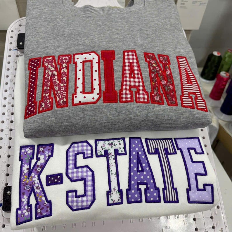 May include: Two sweatshirts, one gray and one white, are stacked. The gray sweatshirt has the word "INDIANA" in red and patterned fabric letters. The white sweatshirt displays "K-STATE" in purple and patterned fabric letters. Both sweatshirts are embroidered.