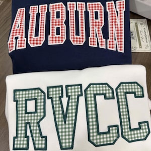 May include: A navy blue sweatshirt with the word AUBURN in red and white gingham applique letters and a white sweatshirt with the word RVCC in green and white gingham applique letters. Both garments feature a classic crew neck design.