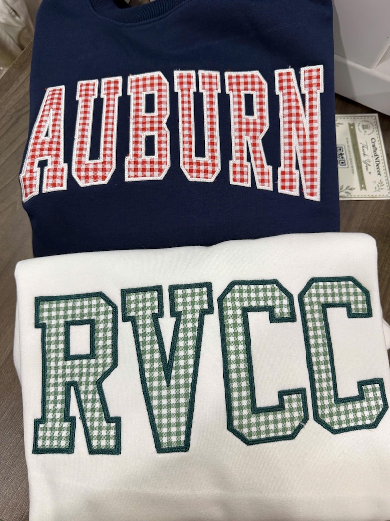 May include: A navy blue sweatshirt with the word AUBURN in red and white gingham applique letters and a white sweatshirt with the word RVCC in green and white gingham applique letters. Both garments feature a classic crew neck design.