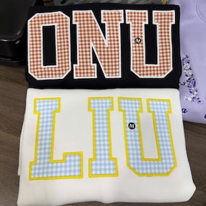 May include: A black sweatshirt featuring the letters ONU in brown and white gingham applique, and a white sweatshirt featuring the letters LIU in light blue and yellow gingham applique. Both garments have a small black size M tag attached.