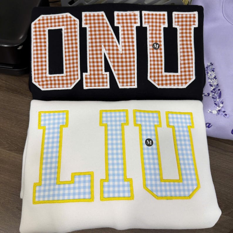 May include: A black sweatshirt featuring the letters ONU in brown and white gingham applique, and a white sweatshirt featuring the letters LIU in light blue and yellow gingham applique. Both garments have a small black size M tag attached.
