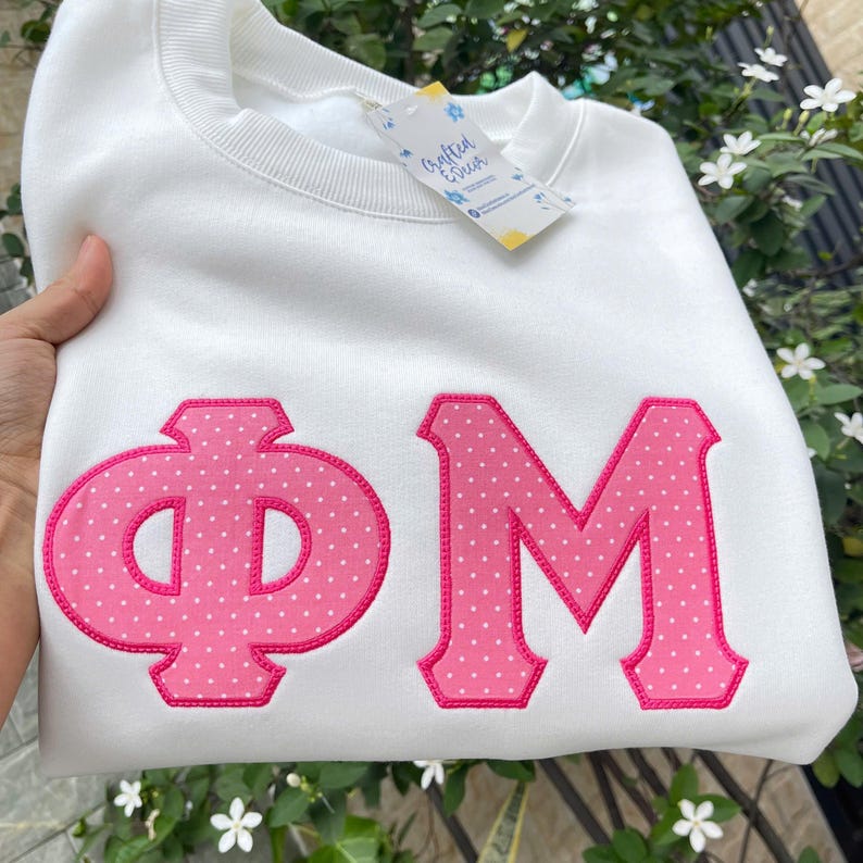 Puede incluir: Sudadera blanca con letras griegas &Phi; y M bordadas en rosa con lunares, contorneadas en rosa m&aacute;s oscuro. Una etiqueta con el texto "Crafted & Deck" est&aacute; adjunta. Prenda de vestir informal.