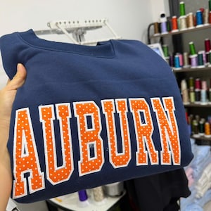 May include: Navy blue sweatshirt with the word "AUBURN" in large orange letters with white polka dots. The sweatshirt is folded, and the text is centered across the chest. The fabric appears to be a soft material.