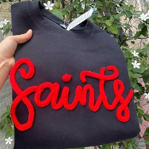 Puede incluir: Una taza de cerámica blanca con asa negra y el texto "But First, Coffee" en negrita negra. La taza se encuentra sobre un fondo borroso, sugiriendo un ambiente acogedor.