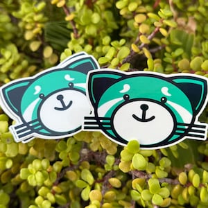May include: Two stickers featuring a cartoon red panda design. The stickers have a white background with a teal and black panda face. The panda has a white face with a black nose and a smiling mouth. The stickers are surrounded by green foliage.