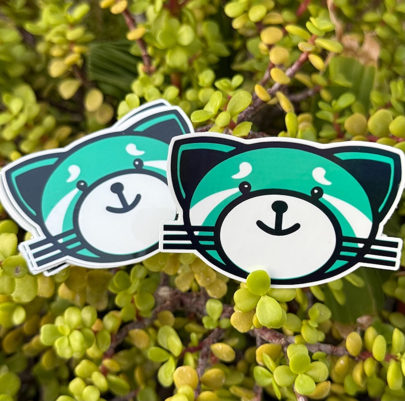 May include: Two stickers featuring a cartoon red panda design. The stickers have a white background with a teal and black panda face. The panda has a white face with a black nose and a smiling mouth. The stickers are surrounded by green foliage.