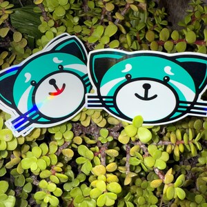 May include: Two stickers featuring a cartoon red panda design. The red panda has a white face, teal accents, and a black outline. The stickers are placed on a background of green foliage.