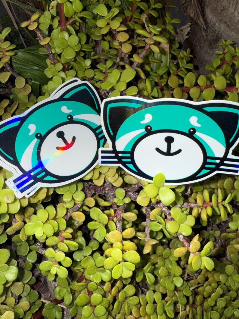 May include: Two stickers featuring a cartoon red panda design. The red panda has a white face, teal accents, and a black outline. The stickers are placed on a background of green foliage.