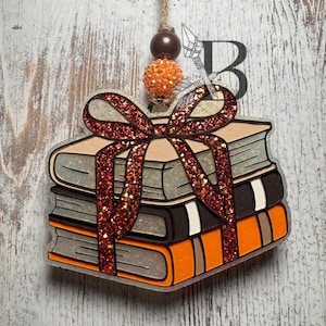 May include: A hanging ornament shaped like a stack of three books tied with a dark red glittery ribbon. The books are colored light gray, black, and orange. A brown wooden bead and an orange glitter bead are attached to the top twine hanger.