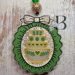 May include: A green and white St. Patrick's Day themed car air freshener. The air freshener has a scalloped green border, a bow, and a design of stacked bowls with shamrocks and hearts. The air freshener is hanging from a black string with green and gold beads.