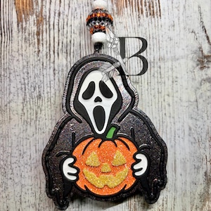 May include: A hanging ornament featuring a black hooded figure with a white ghost mask holding a glittery orange jack-o'-lantern. The ornament is suspended by a black cord with white and orange-black rhinestone-encrusted beads above the figure.