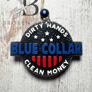 May include: A round, black air freshener with the words "DIRTY HANDS BLUE COLLAR CLEAN MONEY" in white and blue lettering. The design includes red stripes and white stars. It hangs from a string with a blue bead.