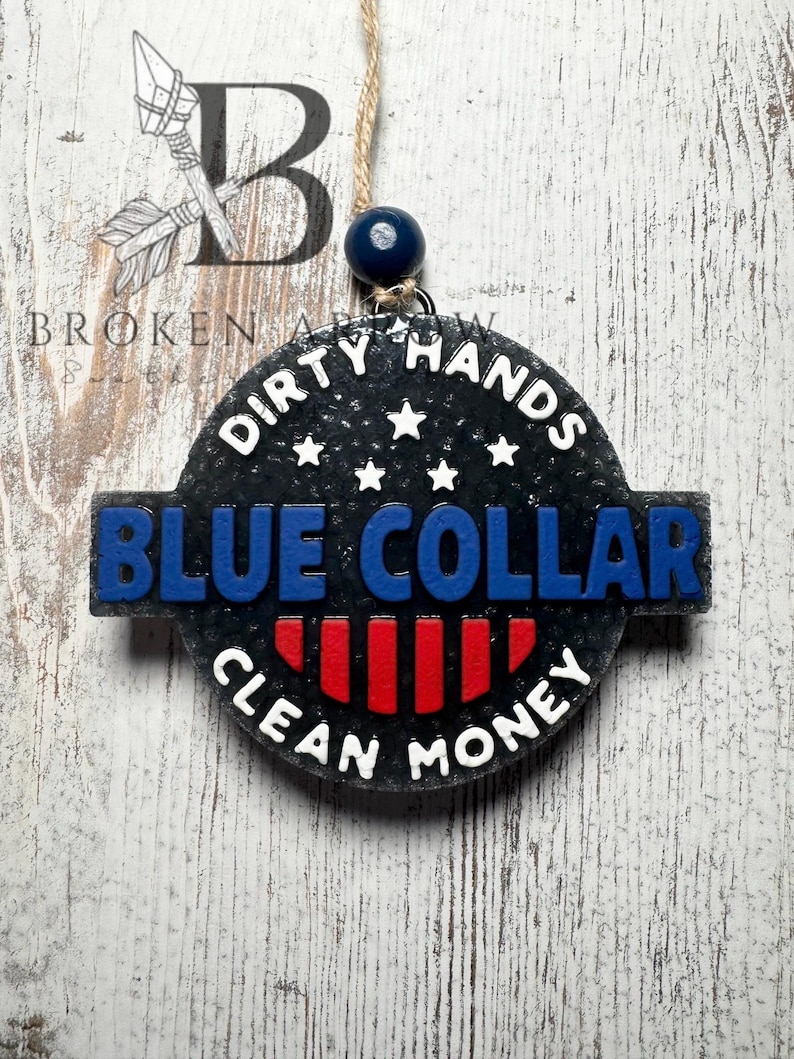 May include: A round, black air freshener with the words "DIRTY HANDS BLUE COLLAR CLEAN MONEY" in white and blue lettering. The design includes red stripes and white stars. It hangs from a string with a blue bead.