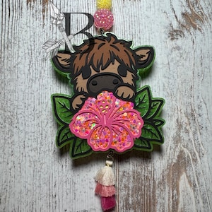 May include: A brown and white Highland cow cartoon charm with a pink glitter hibiscus flower and green leaves. The charm is attached to a pink, white, and cream tassel.