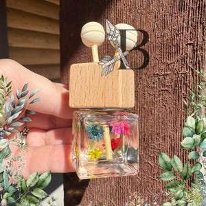 May include: A small, clear glass bottle with a wooden top and a cork stopper. The bottle is filled with dried flowers in shades of pink, yellow, blue, and green. The bottle is a car air freshener.