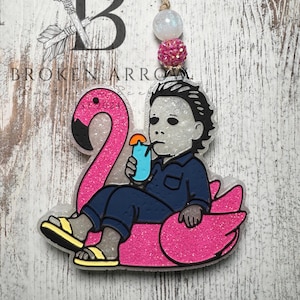 May include: A glittery pink flamingo pool float with a cartoon figure of Michael Myers, holding a blue drink. The figure is wearing a blue jumpsuit and yellow sandals. The image is an air freshener with a decorative string.