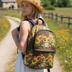 Könnte beinhalten: Ein brauner Rucksack mit einem wiederkehrenden Muster aus gelben Sonnenblumen und grünen Blättern sowie zwei illustrierten Fröschen, die zwischen den Blumen sitzen. Der Rucksack hat ein Hauptfach mit Reißverschluss und eine kleinere Fronttasche.