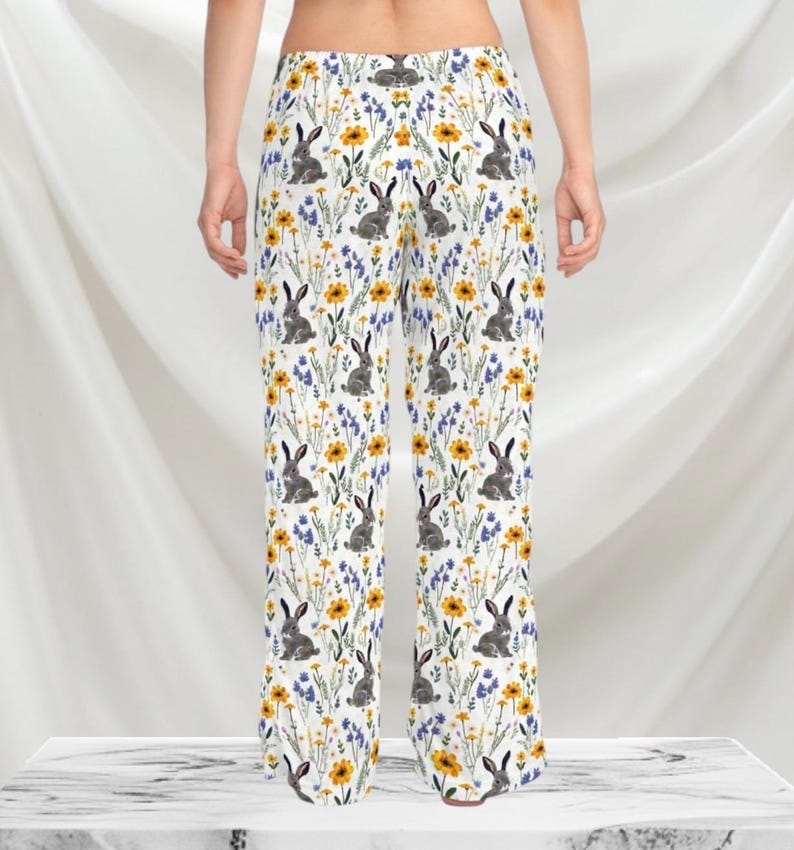 May include: White pajama pants with a repeating pattern of gray rabbits, yellow sunflowers, and blue wildflowers. The pants have a wide leg design and are made of a soft, lightweight fabric.