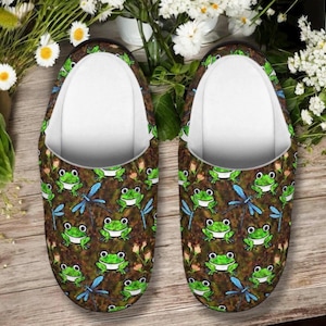 May include: A pair of slip-on slippers featuring a repeating pattern of green frogs, blue dragonflies, and small pink flowers on a dark brown background. The interior of the slippers is lined with a solid white fabric, and the edges are trimmed in black.