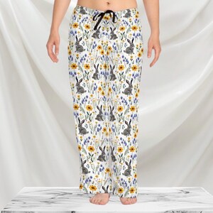 May include: White pajama pants with a repeating pattern of gray rabbits, yellow flowers, and blue wildflowers. The pants have a black drawstring and a relaxed fit. The pajama pants are made of a soft, lightweight fabric.