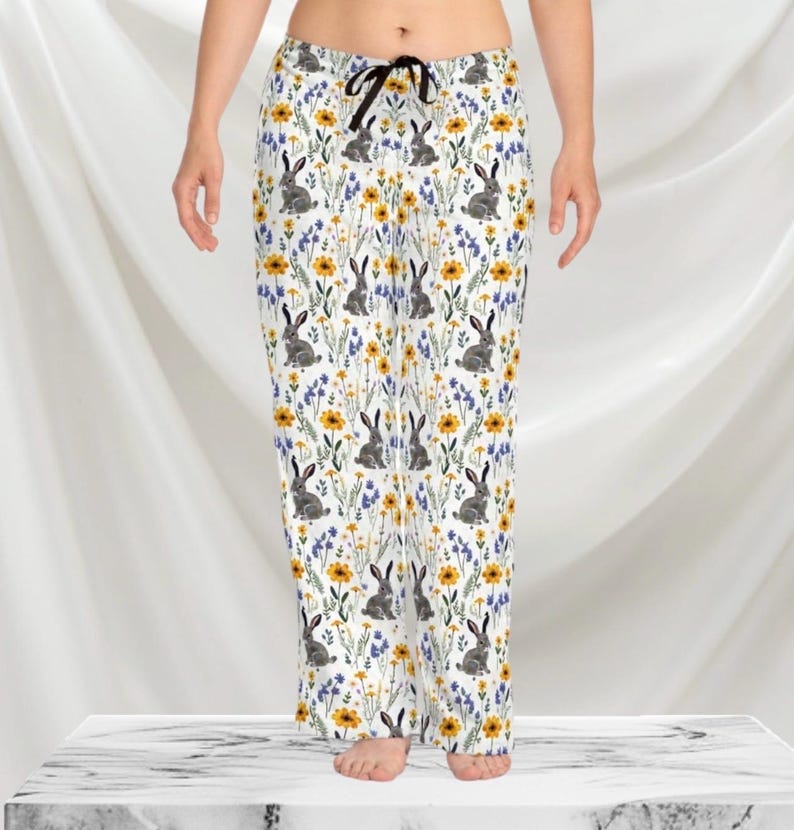 May include: White pajama pants with a repeating pattern of gray rabbits, yellow flowers, and blue wildflowers. The pants have a black drawstring and a relaxed fit. The pajama pants are made of a soft, lightweight fabric.