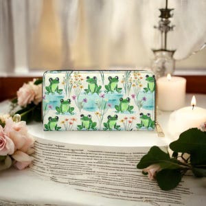 May include: A rectangular wallet with a repeating pattern of green frogs on a light background. The frogs are depicted in a pond setting with flowers and water lilies. The wallet has a gold zipper.