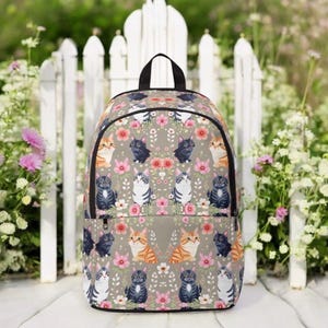 Könnte beinhalten: Ein grauer Rucksack mit einem sich wiederholenden Muster aus Cartoon-Katzen und rosa Blumen. Die Katzen sind in verschiedenen Farben, darunter Orange, Grau und Schwarz. Der Rucksack hat einen schwarzen Griff und eine Vordertasche.