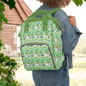 Könnte beinhalten: Ein grüner Rucksack mit einem Froschmuster. Der Rucksack hat ein großes Hauptfach, eine kleinere Fronttasche und verstellbare Träger.