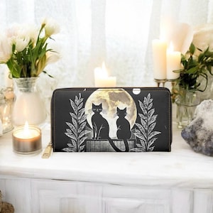 May include: Black zip-around wallet featuring a full moon and two black cat silhouettes. The design includes white botanical accents. The wallet has a gold zipper pull and is set against a white surface with candles and flowers.