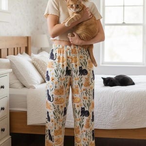 以下が含まれることがあります： クリーム色のリブ編み半袖トップスと、白地に黒とオレンジの猫、黄色い花、緑の葉が全体にプリントされたゆったりとしたパジャマパンツのセット。