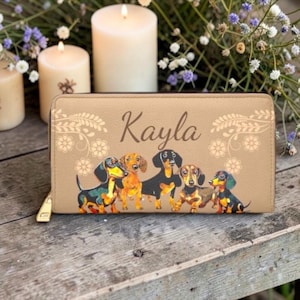 Könnte beinhalten: Beige Geldbörse mit Reißverschluss, verziert mit einer farbenfrohen Illustration von sechs Dackeln. Der Name "Kayla" ist in brauner Schrift über den Hunden aufgedruckt. Florale Muster schmücken die oberen Ecken.