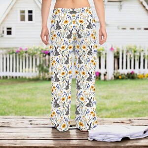 May include: White palazzo pants with a repeating pattern of gray rabbits, yellow sunflowers, and blue wildflowers. The pants have a wide leg and a high waist. The background includes a white picket fence and a house.
