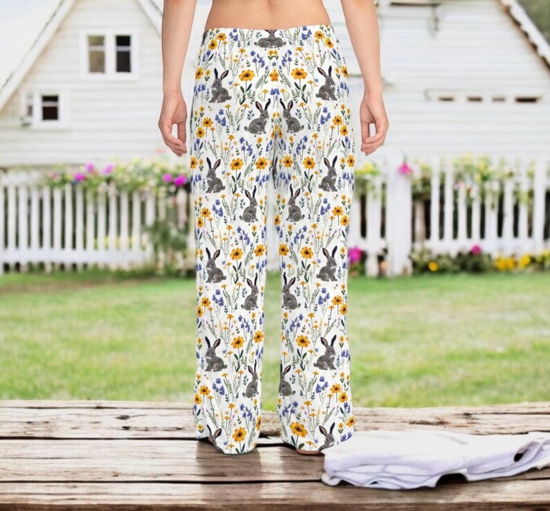 May include: White palazzo pants with a repeating pattern of gray rabbits, yellow sunflowers, and blue wildflowers. The pants have a wide leg and a high waist. The background includes a white picket fence and a house.