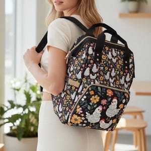 Könnte beinhalten: Schwarzer Rucksack mit einem Muster aus Hühnern und Blumen. Die Tasche hat ein Hauptfach mit Reißverschluss, Vordertaschen und verstellbare Träger. Das Design zeigt weiße Hühner, gelbe und orangefarbene Blumen und grüne Blätter.