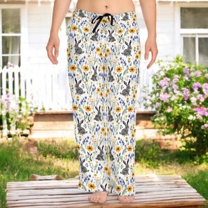 May include: White pajama pants with a repeating pattern of gray rabbits, yellow flowers, and blue wildflowers. The pants have a black drawstring and are made of a soft, lightweight fabric. The overall theme is spring and nature.