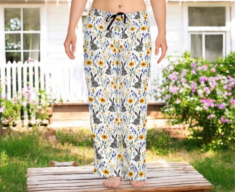 May include: White pajama pants with a repeating pattern of gray rabbits, yellow flowers, and blue wildflowers. The pants have a black drawstring and are made of a soft, lightweight fabric. The overall theme is spring and nature.