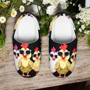 May include: A pair of black house slippers featuring a repeating pattern of cartoon chickens wearing sunglasses. The chickens are yellow, white, and pink with red combs. The slippers have a white interior and are set against a wooden surface.