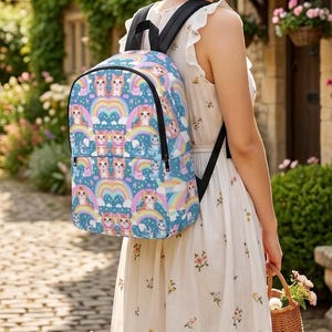 Könnte beinhalten: Ein blauer Rucksack mit einem sich wiederholenden Muster aus orangefarbenen getigerten Katzen, bunten Regenbögen und kleinen weißen Wolken. Der Rucksack verfügt über schwarze Träger, ein Hauptfach mit Reißverschluss und eine Vordertasche.