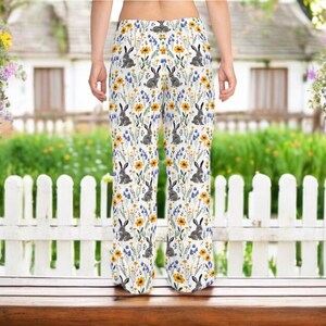 May include: White pajama pants with a repeating pattern of gray rabbits, yellow sunflowers, and blue wildflowers. The pants are wide-legged and made of a lightweight fabric, perfect for lounging. The background shows a white picket fence and a house.