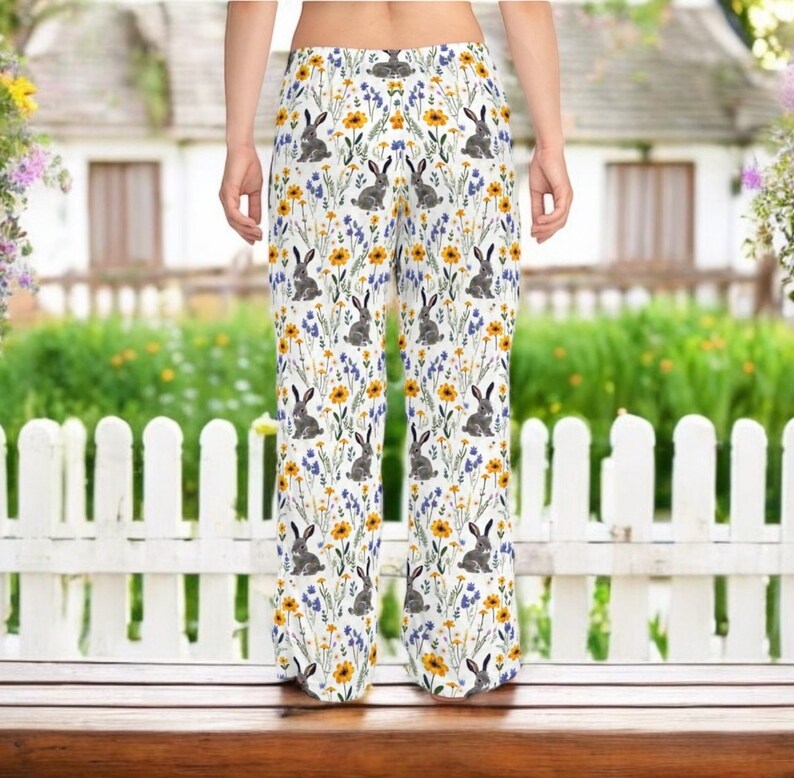 May include: White pajama pants with a repeating pattern of gray rabbits, yellow sunflowers, and blue wildflowers. The pants are wide-legged and made of a lightweight fabric, perfect for lounging. The background shows a white picket fence and a house.