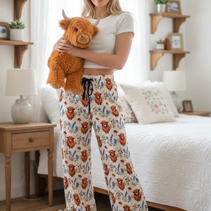 May include: Cream-colored pajama pants with a repeating pattern of Highland cows, blue flowers, and red flowers. The pants have a drawstring waist. A plush Highland cow toy is held in the image.