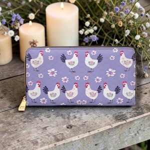 May include: A rectangular zip-around wallet in a light purple color featuring a repeating pattern of white chickens with red combs and small white flowers. The wallet has a gold-toned metal zipper pull and a quilted texture across the surface.