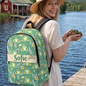 May include: A green backpack featuring a repeating pattern of cartoon frogs and yellow flowers on a green background. A horizontal cream-colored band across the center displays the name Sofie in dark green script. The bag has black straps.
