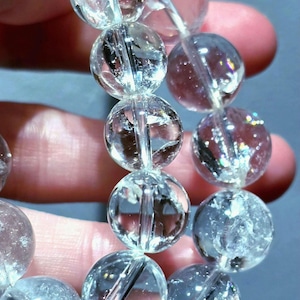 Può includere: Un braccialetto di perline di cristallo di quarzo trasparente. Le perline sferiche sono trasparenti e infilate su un cordino elastico trasparente. Il braccialetto è mostrato su uno sfondo blu, evidenziando la chiarezza e la brillantezza del quarzo.