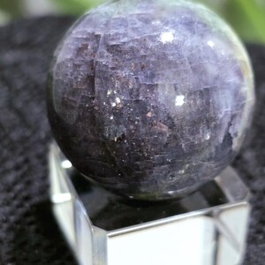 K&ouml;nnte beinhalten: Eine dunkelviolette Kugel mit einem funkelnden, irisierenden Finish. Die Kugel steht auf einem klaren Glasst&auml;nder.