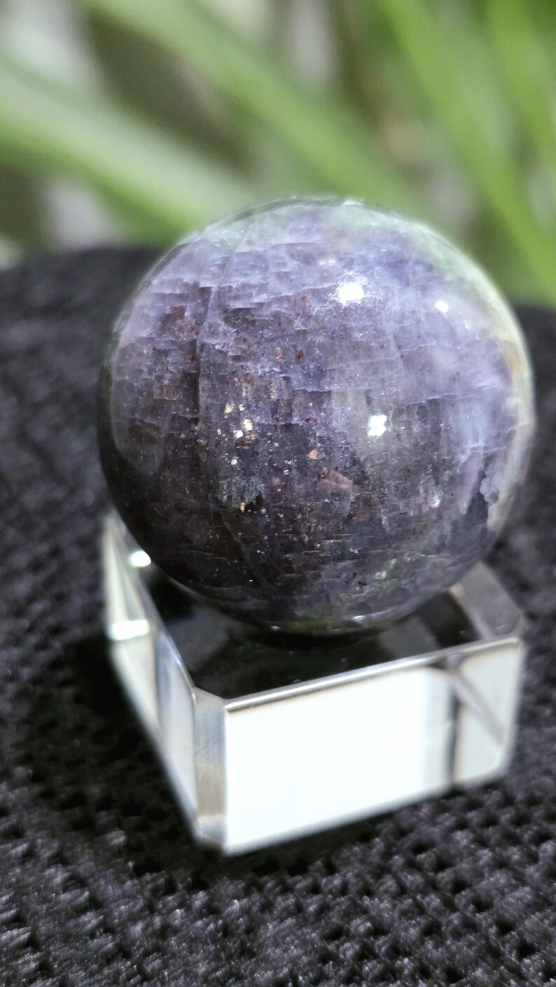 K&ouml;nnte beinhalten: Eine dunkelviolette Kugel mit einem funkelnden, irisierenden Finish. Die Kugel steht auf einem klaren Glasst&auml;nder.