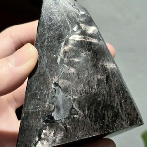 May include: A dark grey, triangular crystal with a polished surface. The crystal has streaks of white and silver. The shape is a pyramid, and it is held in a hand.