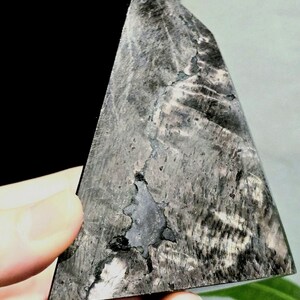 May include: A triangular, polished stone in shades of grey and black. The stone features a distinctive pattern of lines and textures, with a dark, irregular area at its centre. The edges are smooth, and the overall shape is geometric.