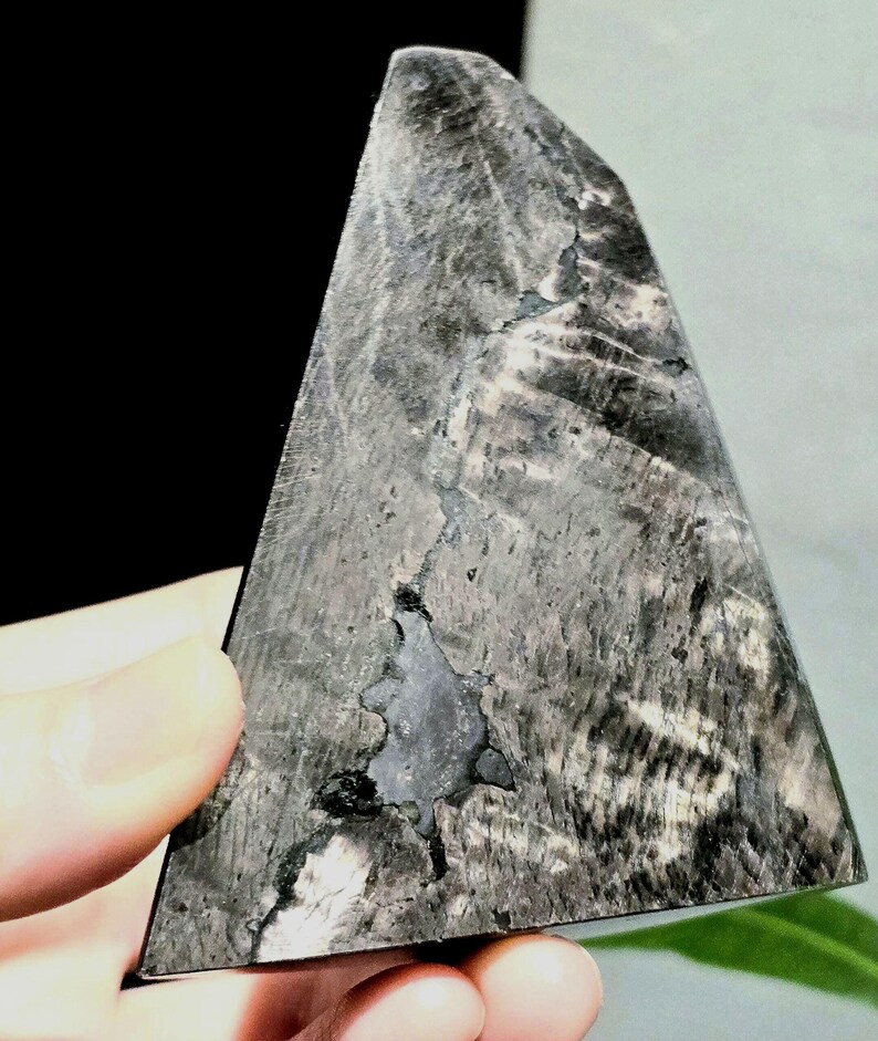 May include: A triangular, polished stone in shades of grey and black. The stone features a distinctive pattern of lines and textures, with a dark, irregular area at its centre. The edges are smooth, and the overall shape is geometric.